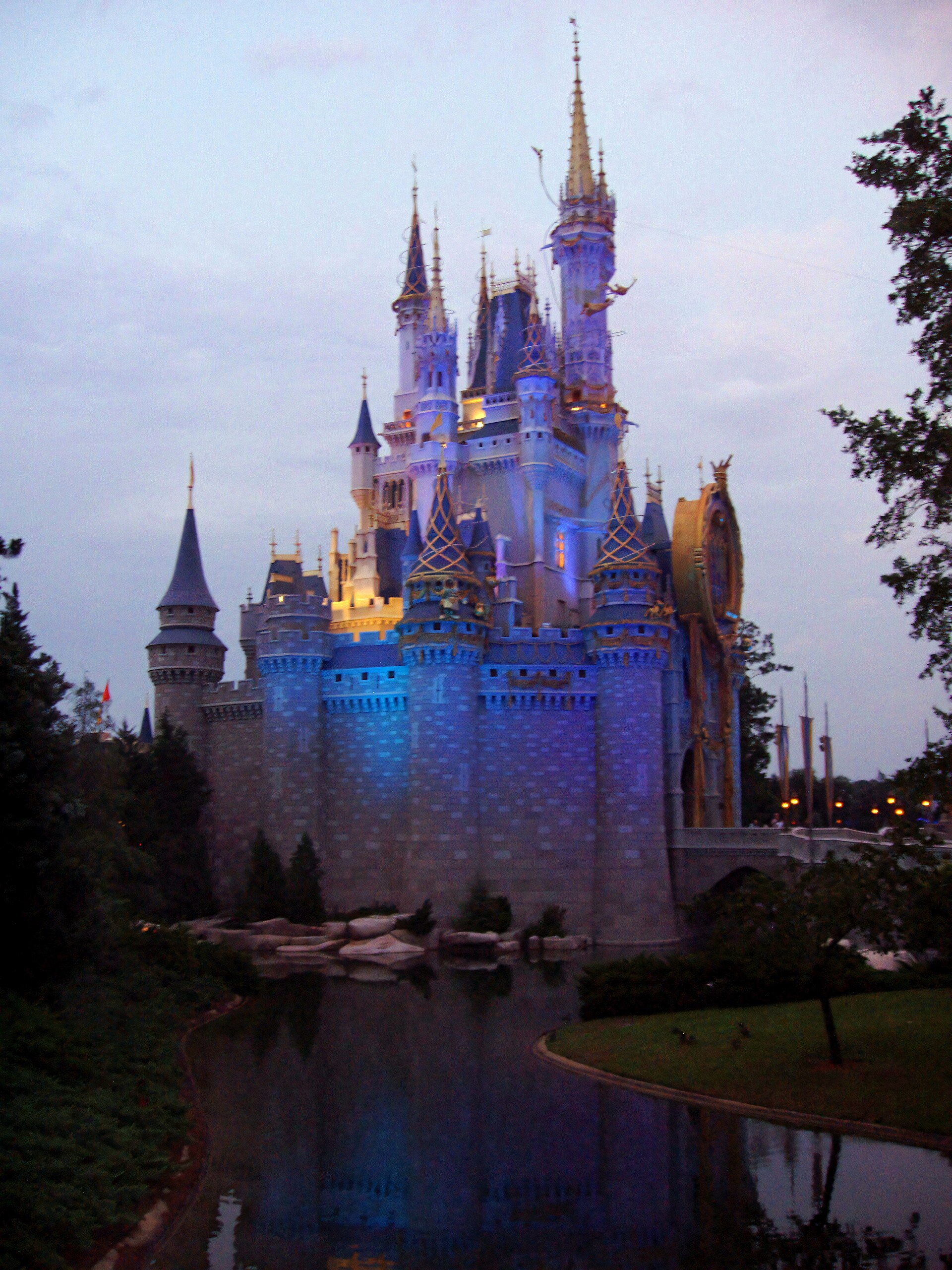 Disney After Hours: el castillo de Cenicienta de Magic Kingdom iluminado de azul y morado al anochecer, reflejado en el agua entre árboles