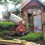 Estatura mínima en Disney: fila de The Barnstormer con un gran cañón blanco de Goofy, carteles de circo de colores y un granero rojo de madera entre plantas