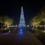 Festival of the Holidays en EPCOT: alto árbol de Navidad con luces azules y guirnaldas doradas brilla de noche al final de un paseo con árboles y visitantes