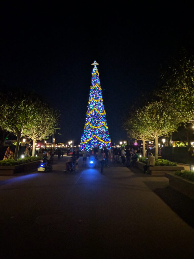 Festival of the Holidays en EPCOT: alto árbol de Navidad con luces azules y guirnaldas doradas brilla de noche al final de un paseo con árboles y visitantes