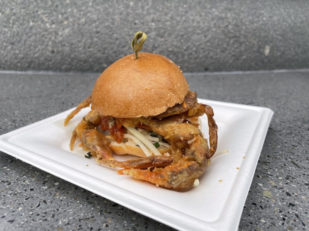 Festival of the Arts: un pequeño sándwich de cangrejo de caparazón blando con pan dorado y un palillo, servido en un plato blanco sobre una mesa gris