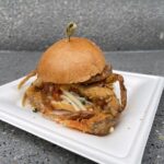 Festival of the Arts: un pequeño sándwich de cangrejo de caparazón blando con pan dorado y un palillo, servido en un plato blanco sobre una mesa gris