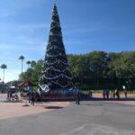 Hollywood Studios en Navidad, parque de Jollywood Nights: alto árbol decorado con guirnaldas plateadas y esferas en una plaza con visitantes y palmeras
