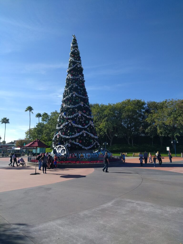 Hollywood Studios en Navidad, parque de Jollywood Nights: alto árbol decorado con guirnaldas plateadas y esferas en una plaza con visitantes y palmeras