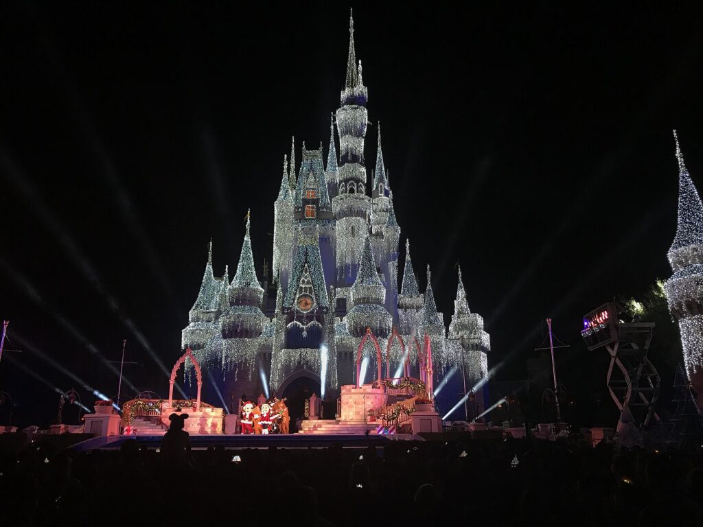 Very Merry Christmas Party: el castillo de Cenicienta cubierto de luces blancas de noche, con personajes vestidos de Navidad en un escenario y público delante