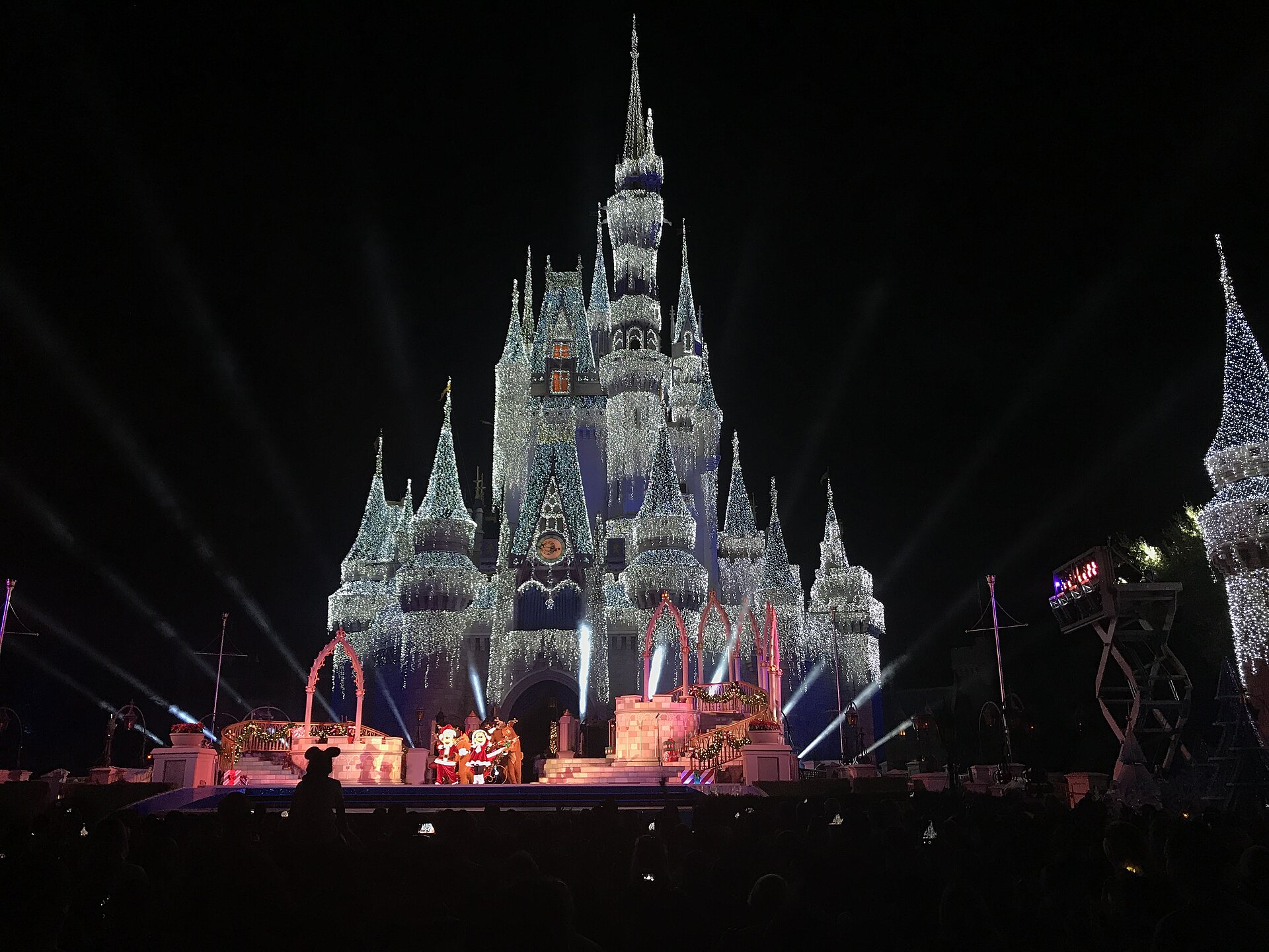 Very Merry Christmas Party: el castillo de Cenicienta cubierto de luces blancas de noche, con personajes vestidos de Navidad en un escenario y público delante