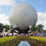 Disney PhotoPass: la gran esfera plateada de Spaceship Earth en EPCOT, con visitantes caminando delante y jardines de flores amarillas bajo un cielo azul con nubes