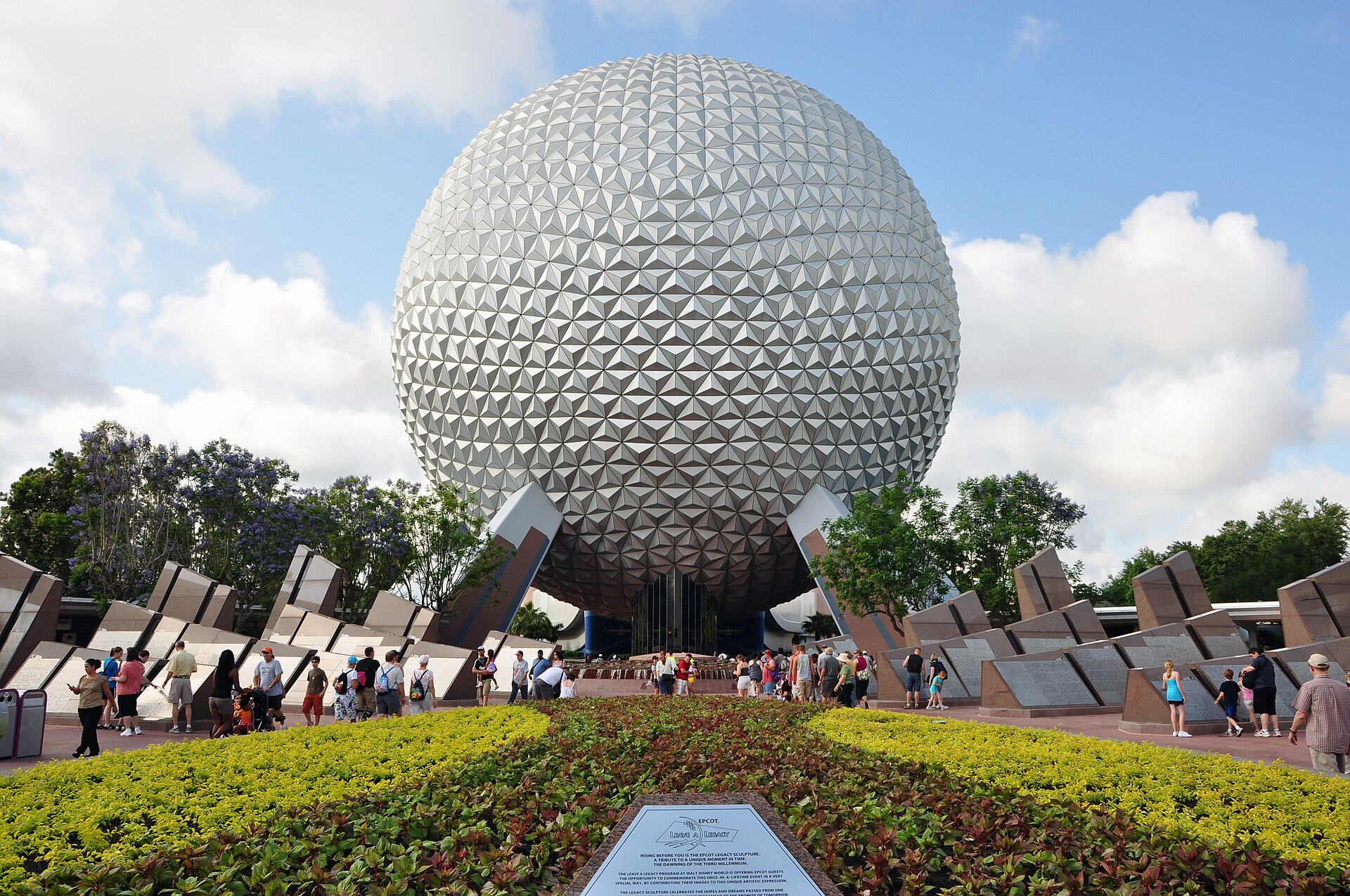 Disney PhotoPass: la gran esfera plateada de Spaceship Earth en EPCOT, con visitantes caminando delante y jardines de flores amarillas bajo un cielo azul con nubes
