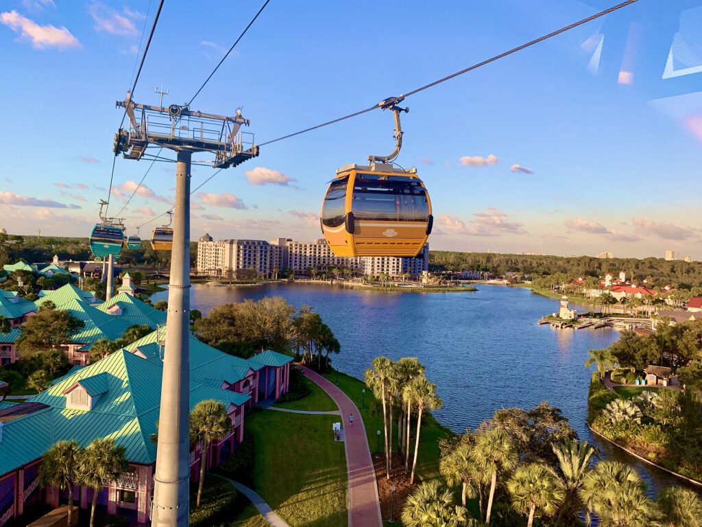 Moverse en Disney Orlando: cabinas del Disney Skyliner colgadas de cables sobre un lago, con palmeras, hoteles de techos verdes y edificios al fondo