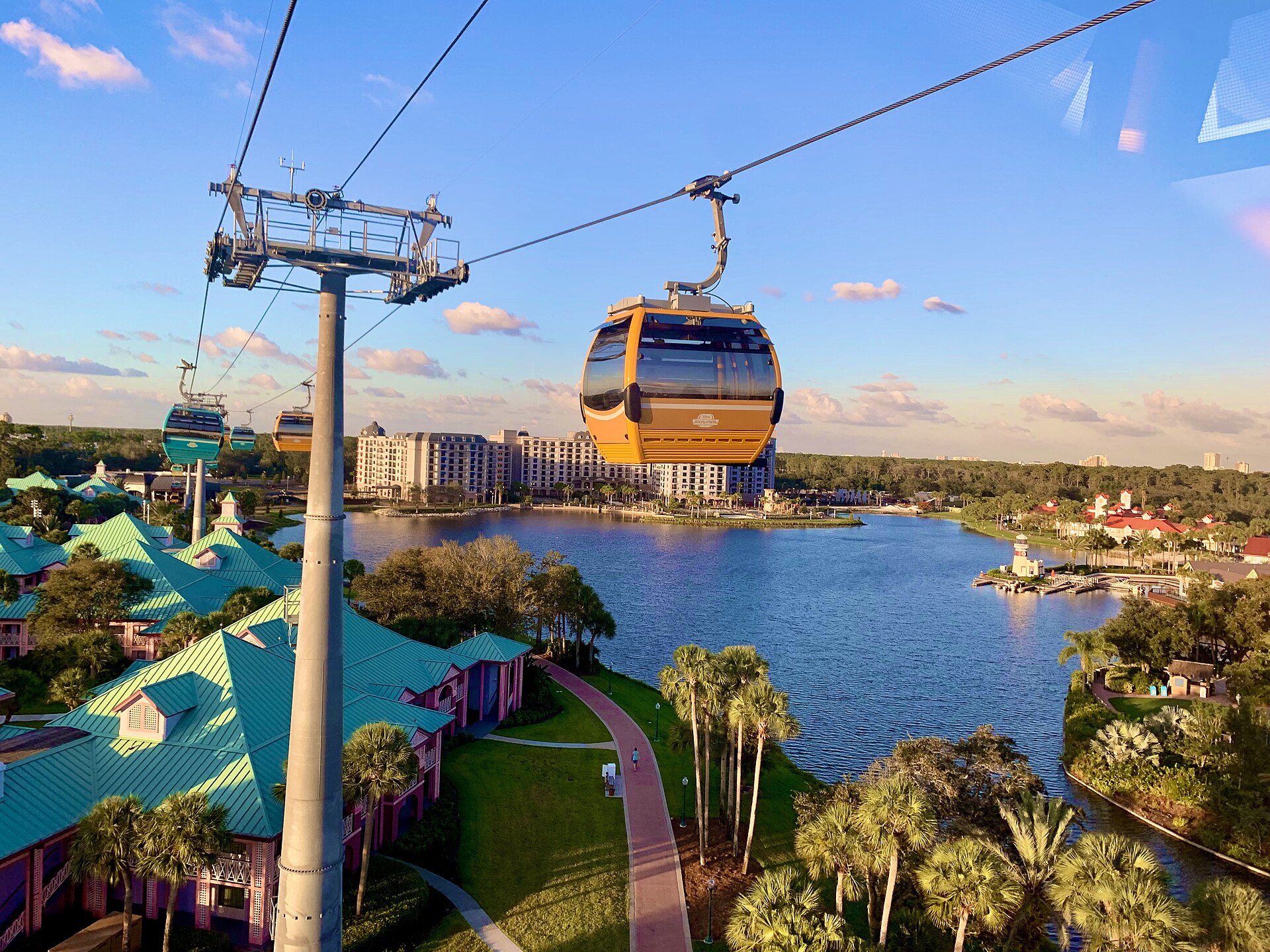 Moverse en Disney Orlando: cabinas del Disney Skyliner colgadas de cables sobre un lago, con palmeras, hoteles de techos verdes y edificios al fondo