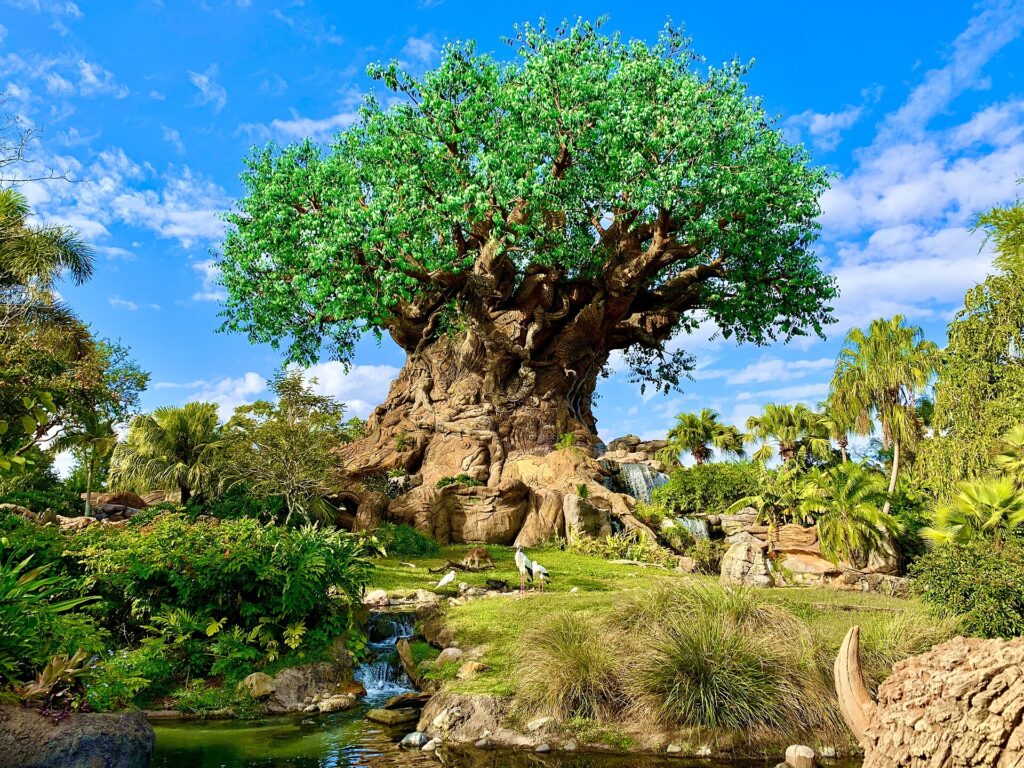 Animal Kingdom: el Tree of Life, un árbol enorme de copa verde, junto a un arroyo, rocas y palmeras, bajo un cielo azul con nubes